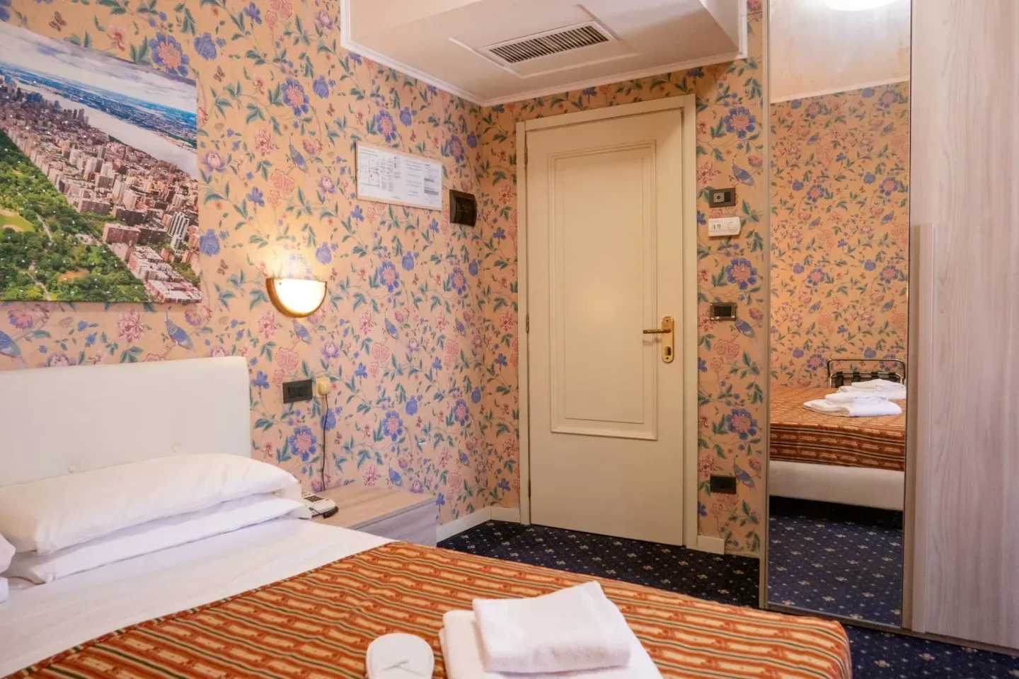 Hotel room with floral wallpaper, a bed, a closed door, and a mirror reflecting the room.