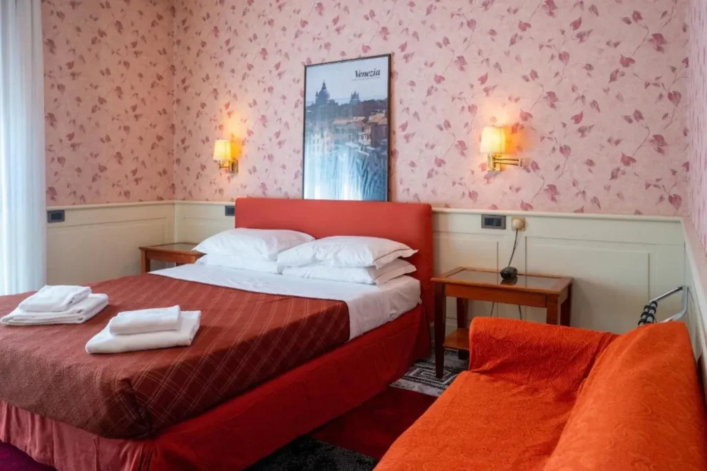 Cozy hotel room with a red bed, sofa, and wallpaper with a floral pattern. A cityscape print hangs above the bed.