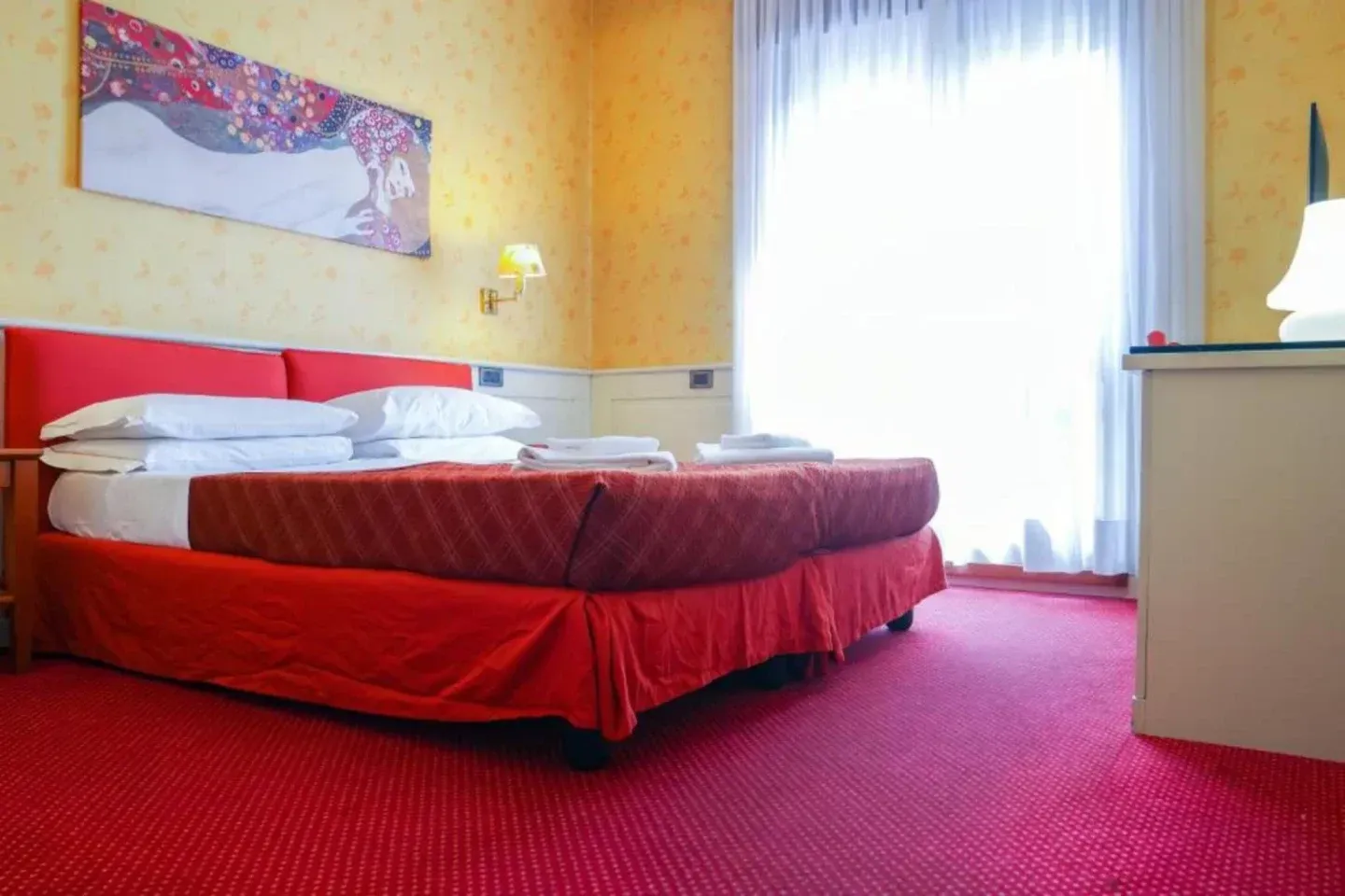 Red-themed hotel room with a bed, red carpet, and bright window. Artwork on wall, nightstand with lamp.