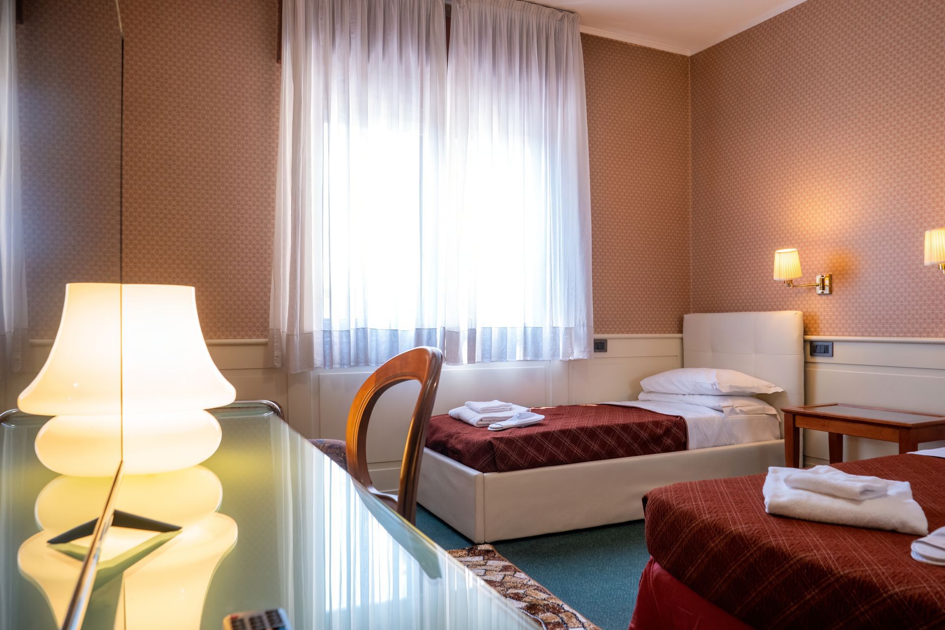 A hotel room with two twin beds, a desk with a lamp, and a window with sheer curtains.