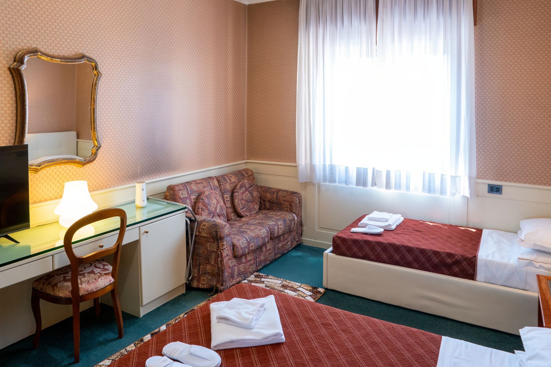 Hotel room with two beds, a small sofa, desk, mirror, and a window with sheer curtains. The room has patterned wallpaper.