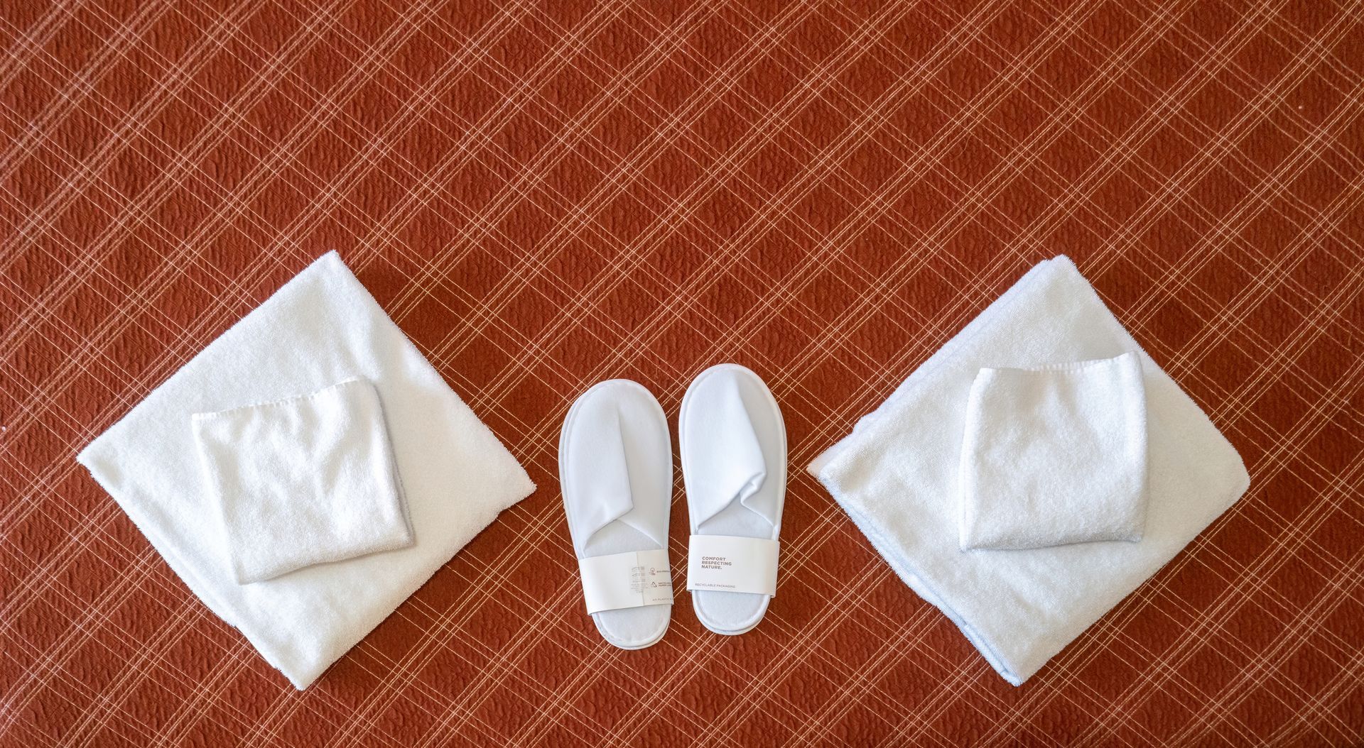 White towels and slippers neatly arranged on a patterned red-orange surface, possibly a hotel bed.