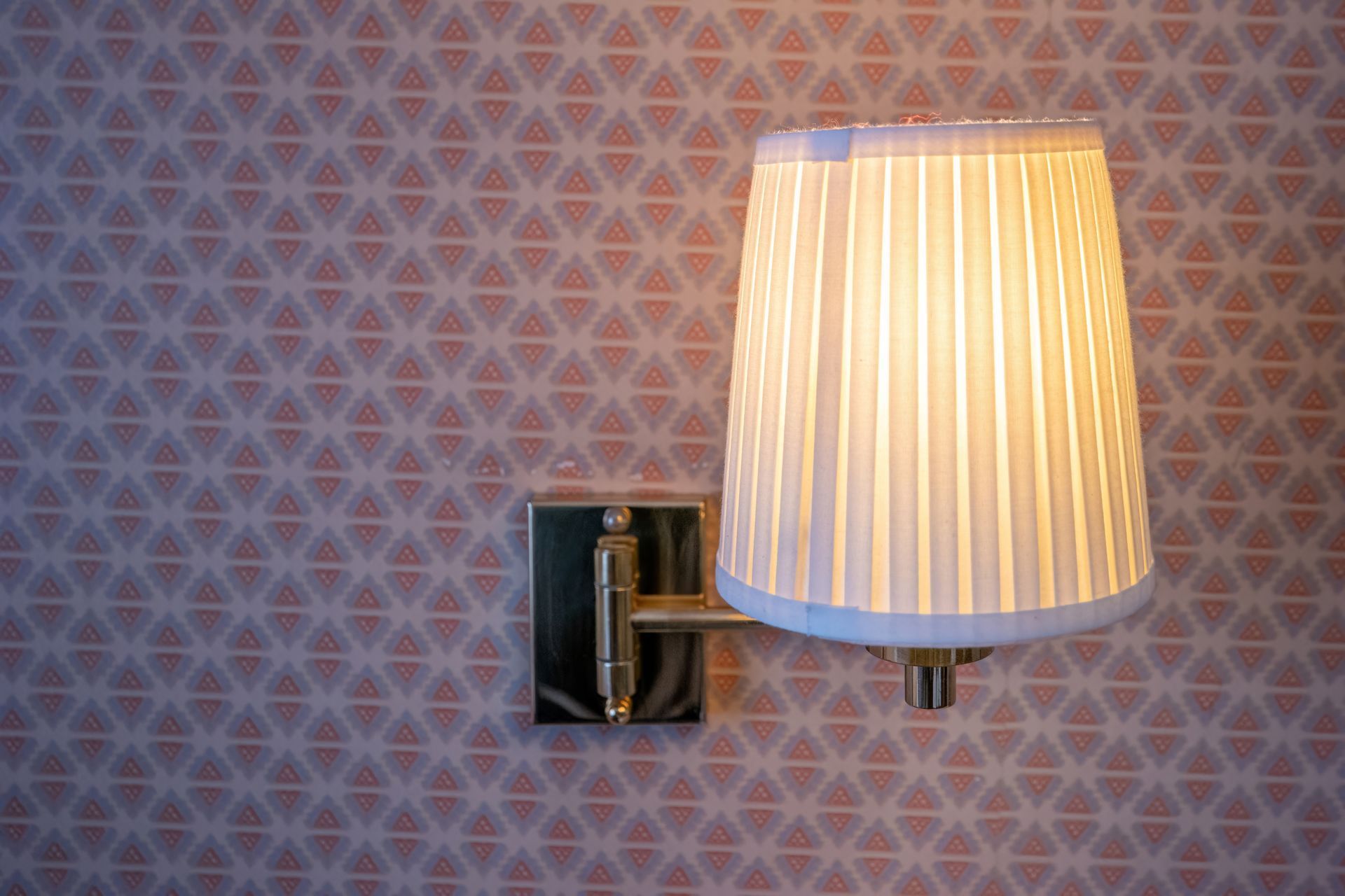 A lit wall sconce with a pleated lampshade against a patterned pink wallpaper.