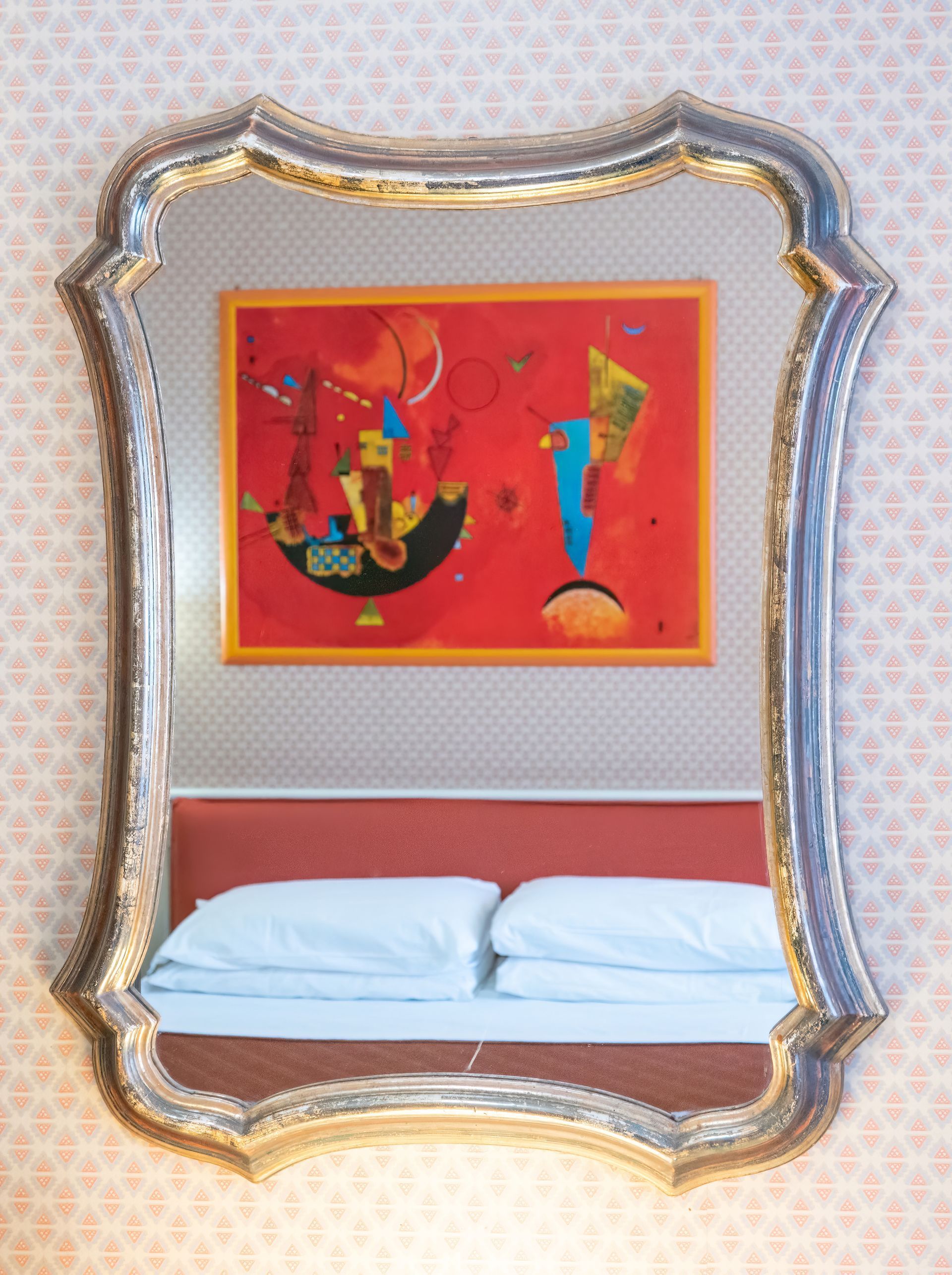 Mirror reflecting a bed with white pillows and an abstract red painting on a pink patterned wall.