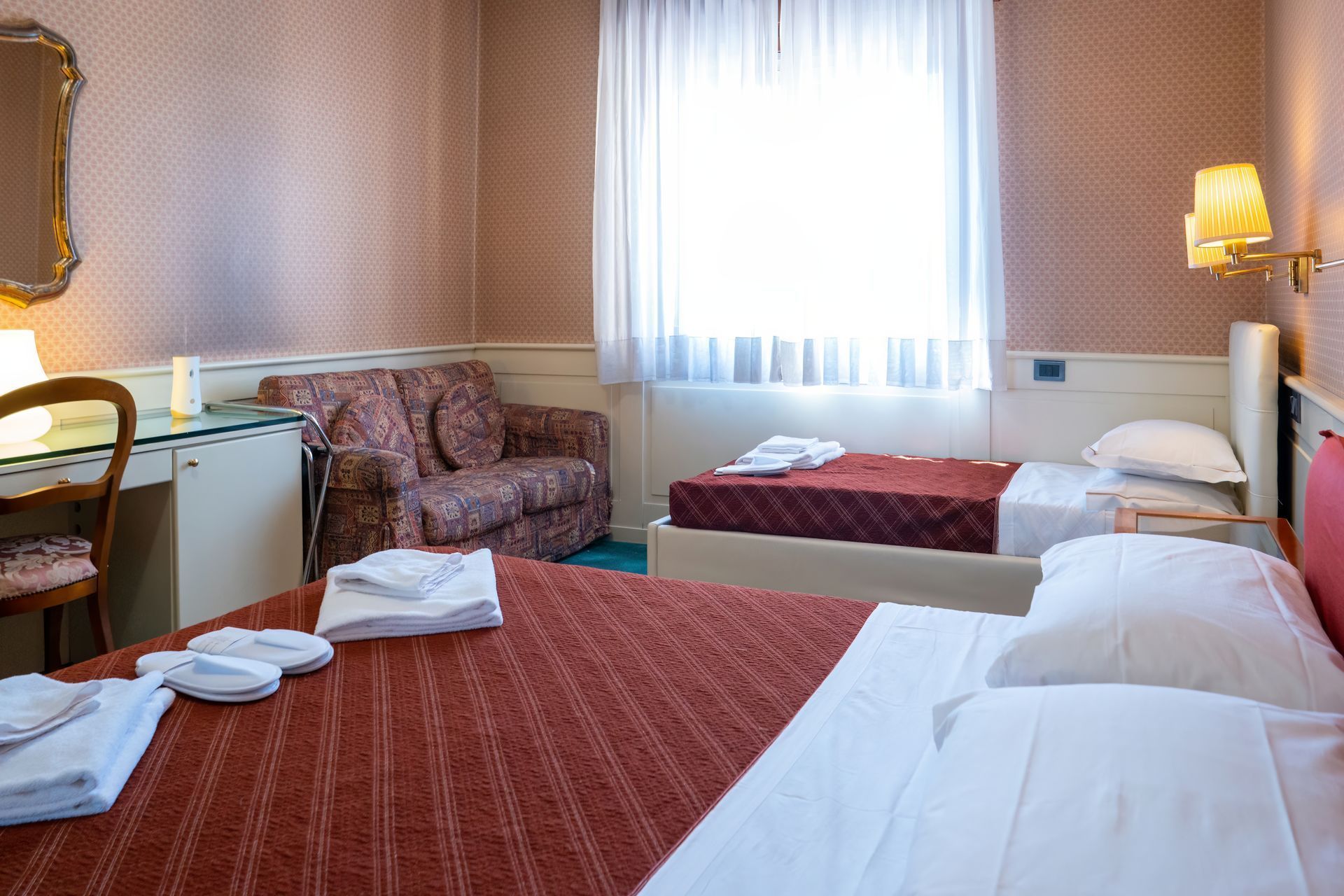 Hotel room with a bed, a single bed, a sofa, and a desk near a window with white curtains. Pink wallpaper.