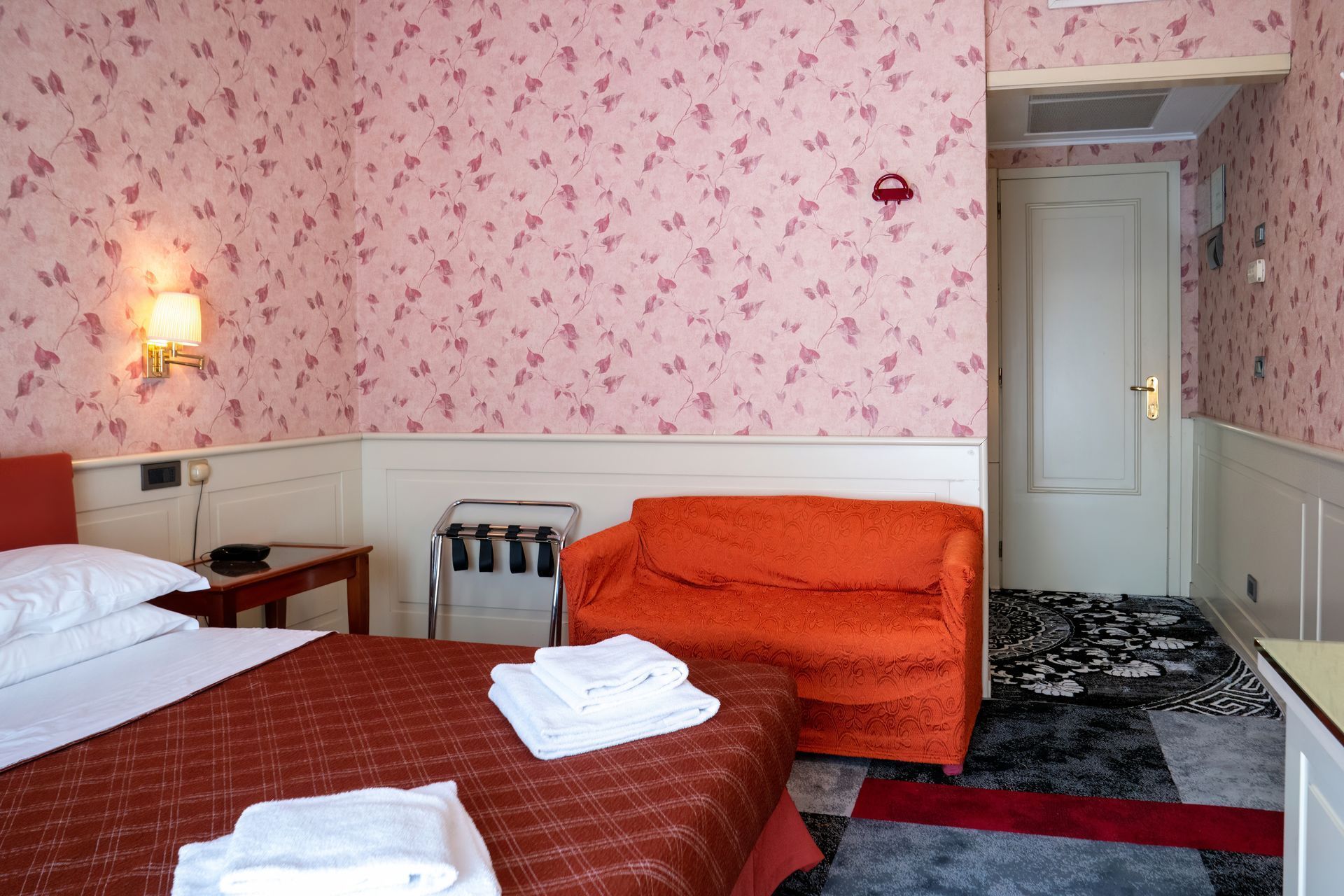 Hotel room with a bed, orange sofa, patterned wallpaper, and a door. There is a luggage rack and a small table.