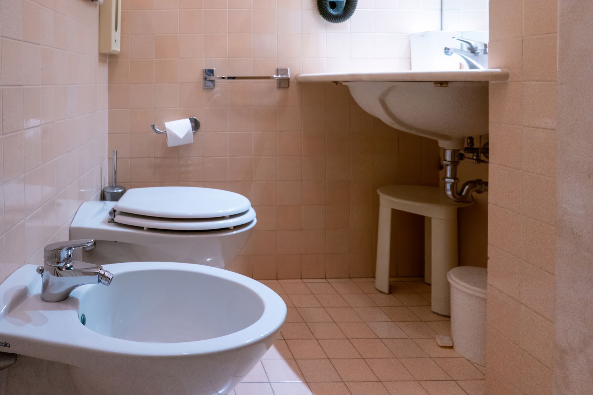 A small bathroom with peach-colored tile walls, including a toilet, bidet, sink with a stool, and a wastebasket.