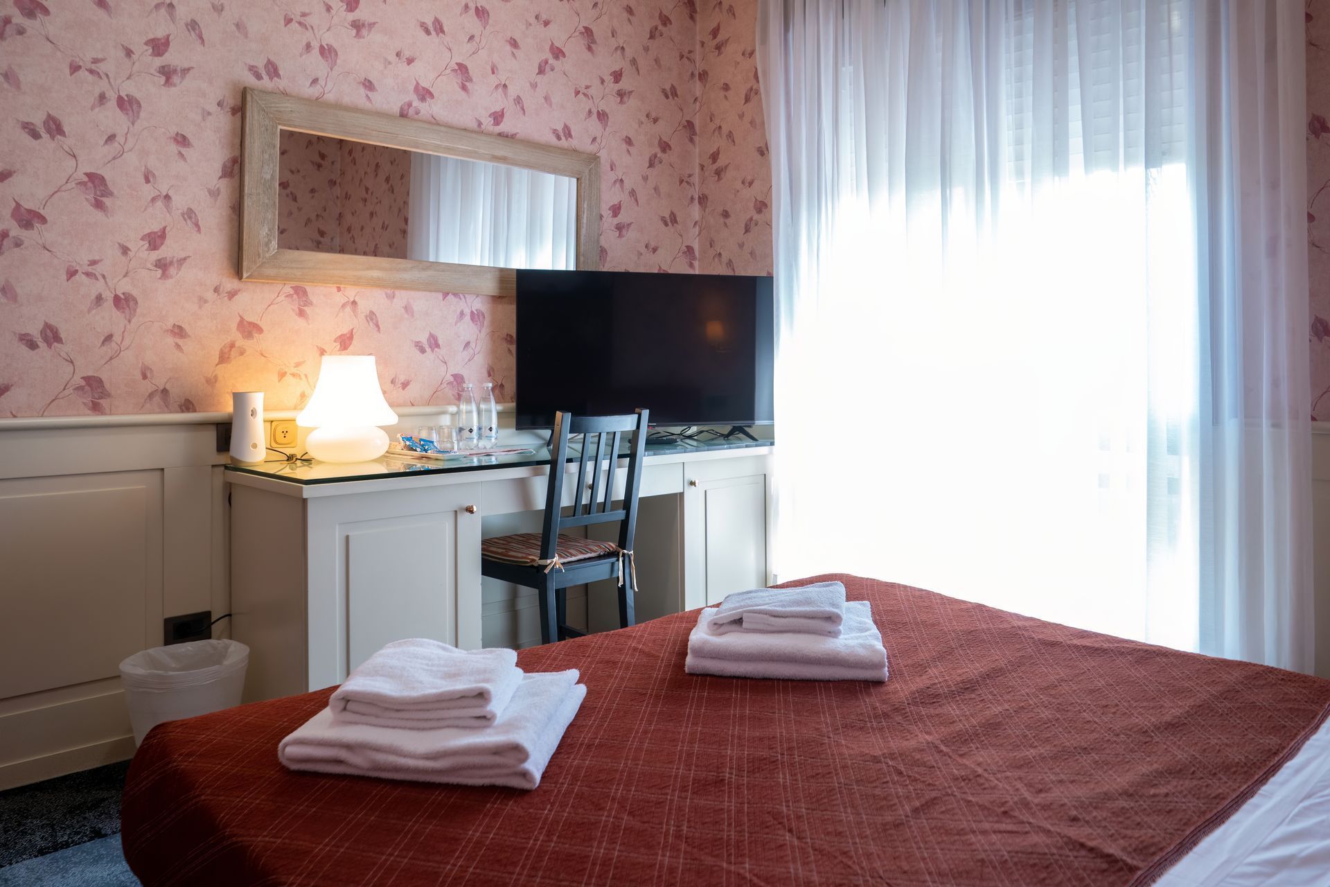Bedroom with a red bed, white towels, a desk with a TV, and a bright window with sheer curtains. 