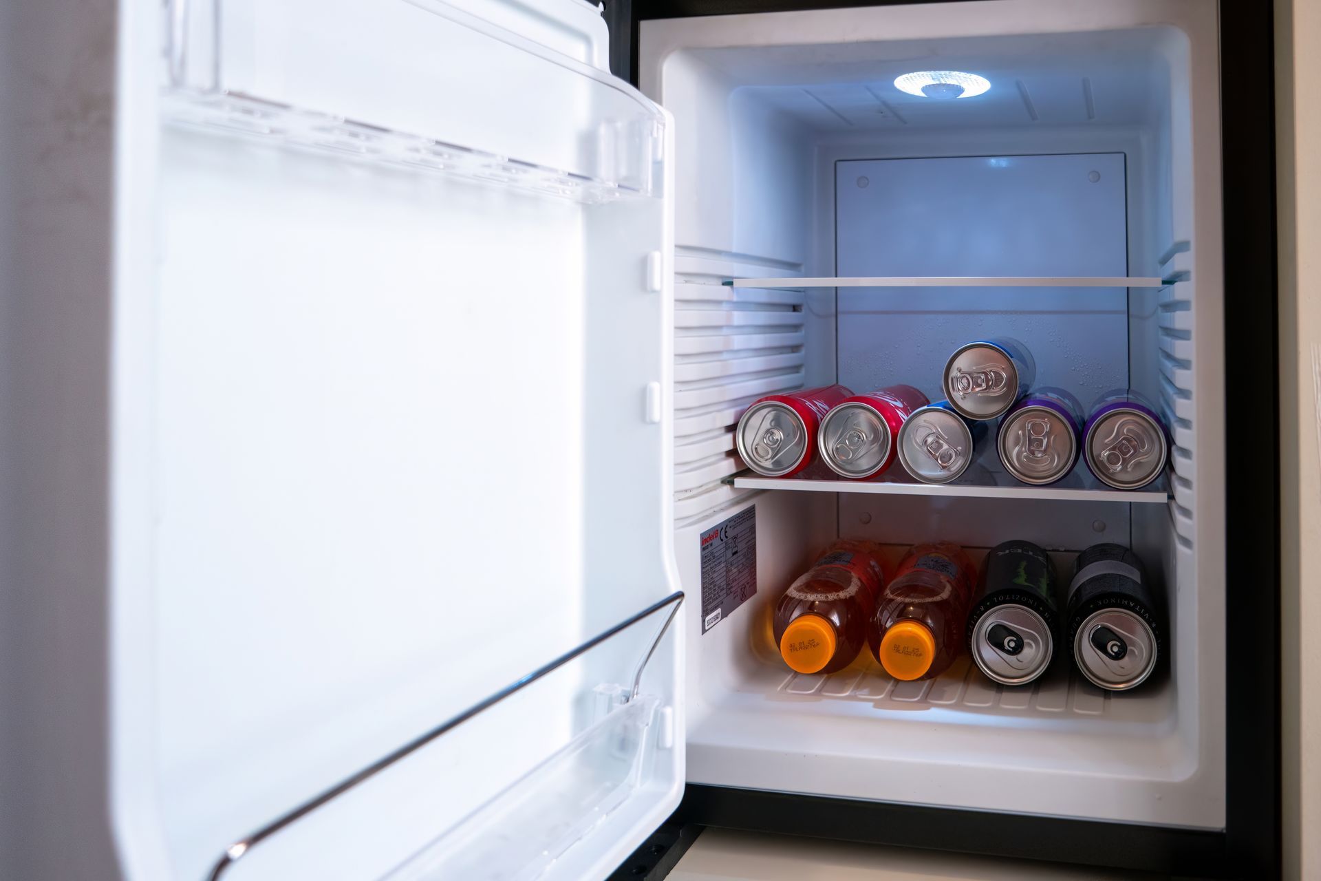 A small, open mini-fridge with cans of soda and juice bottles inside. The fridge is illuminated by an interior light.