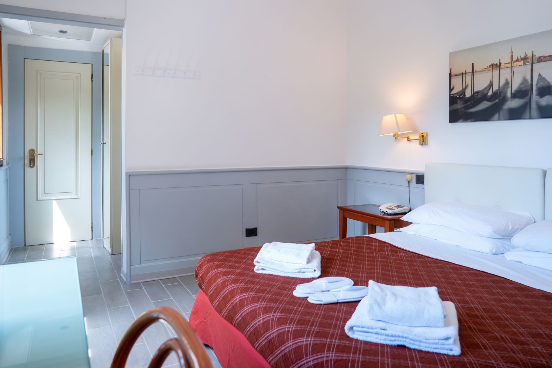Hotel room with a made bed, white towels, and a door leading outside. Painting of gondolas on the wall.