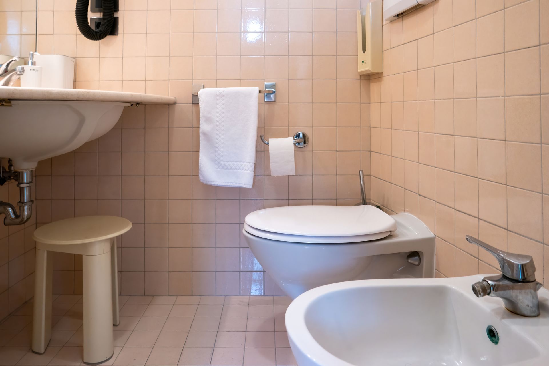 A small bathroom with a sink, toilet, bidet, and a towel rack. The walls and floor are tiled in a light pink color.