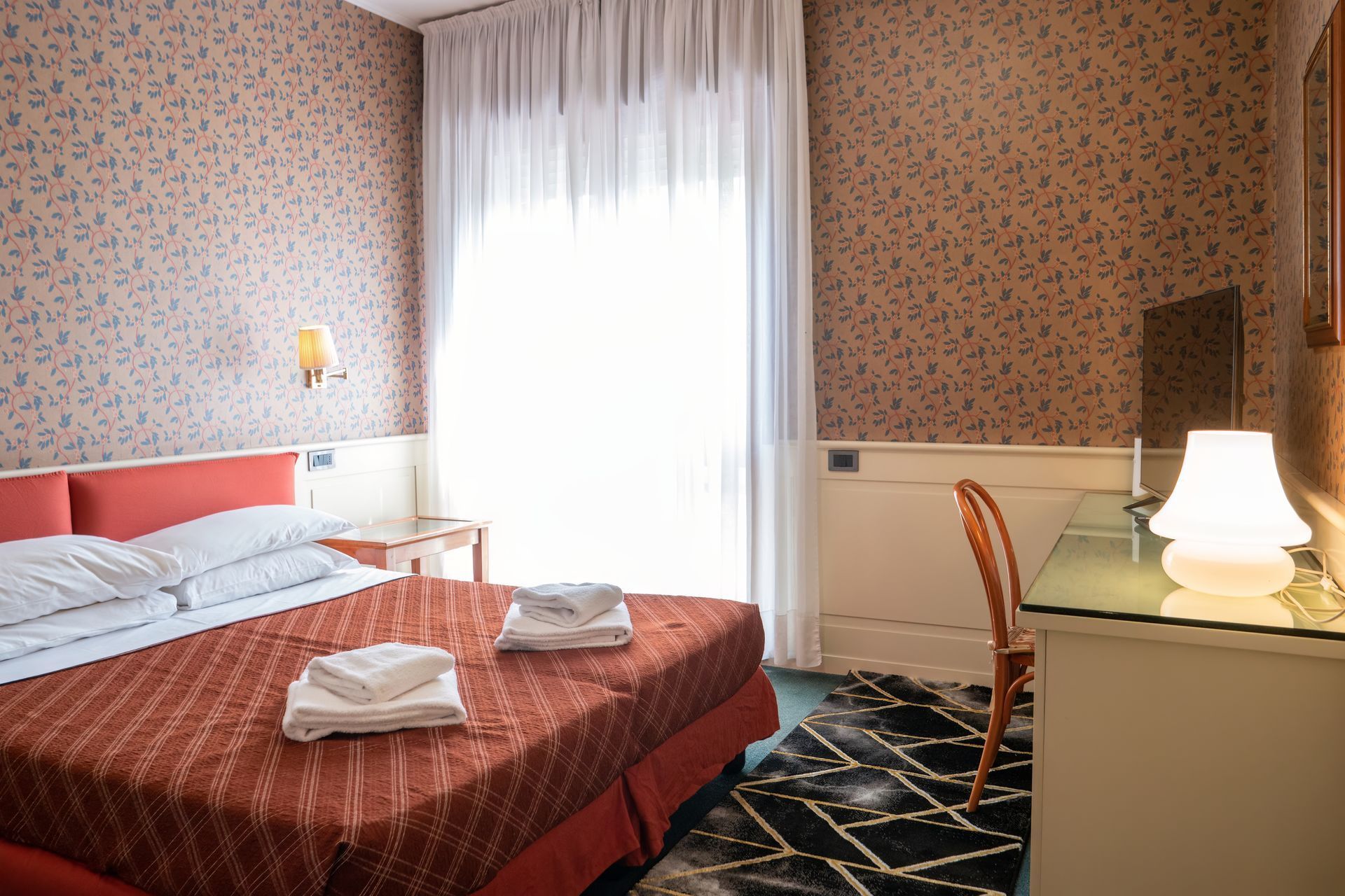 Hotel room with a red bed, desk, and window with sheer curtains. Patterned wallpaper and a patterned rug are also visible.