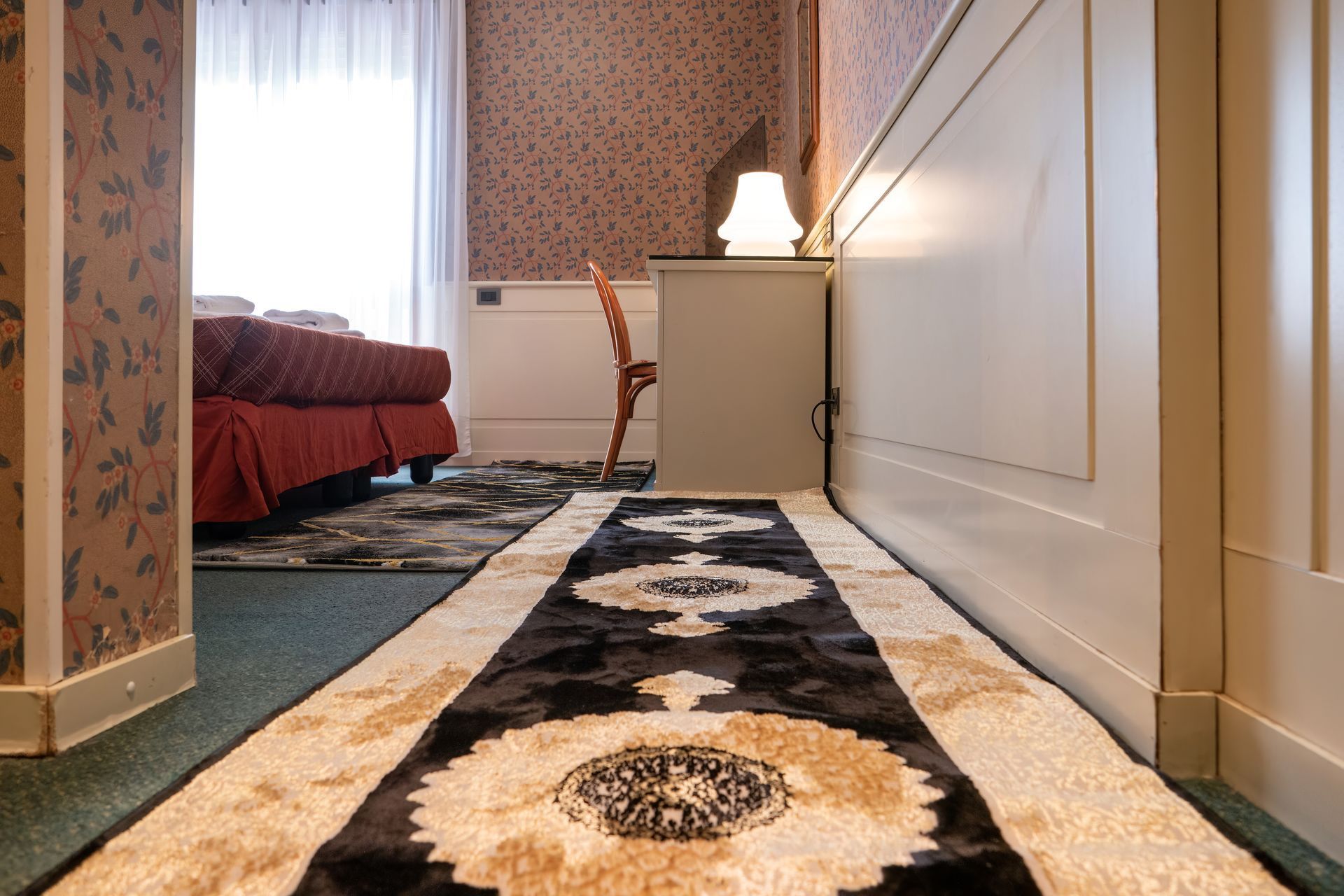 Long hallway with a patterned rug, leading to a bed in a room with patterned wallpaper and a desk.