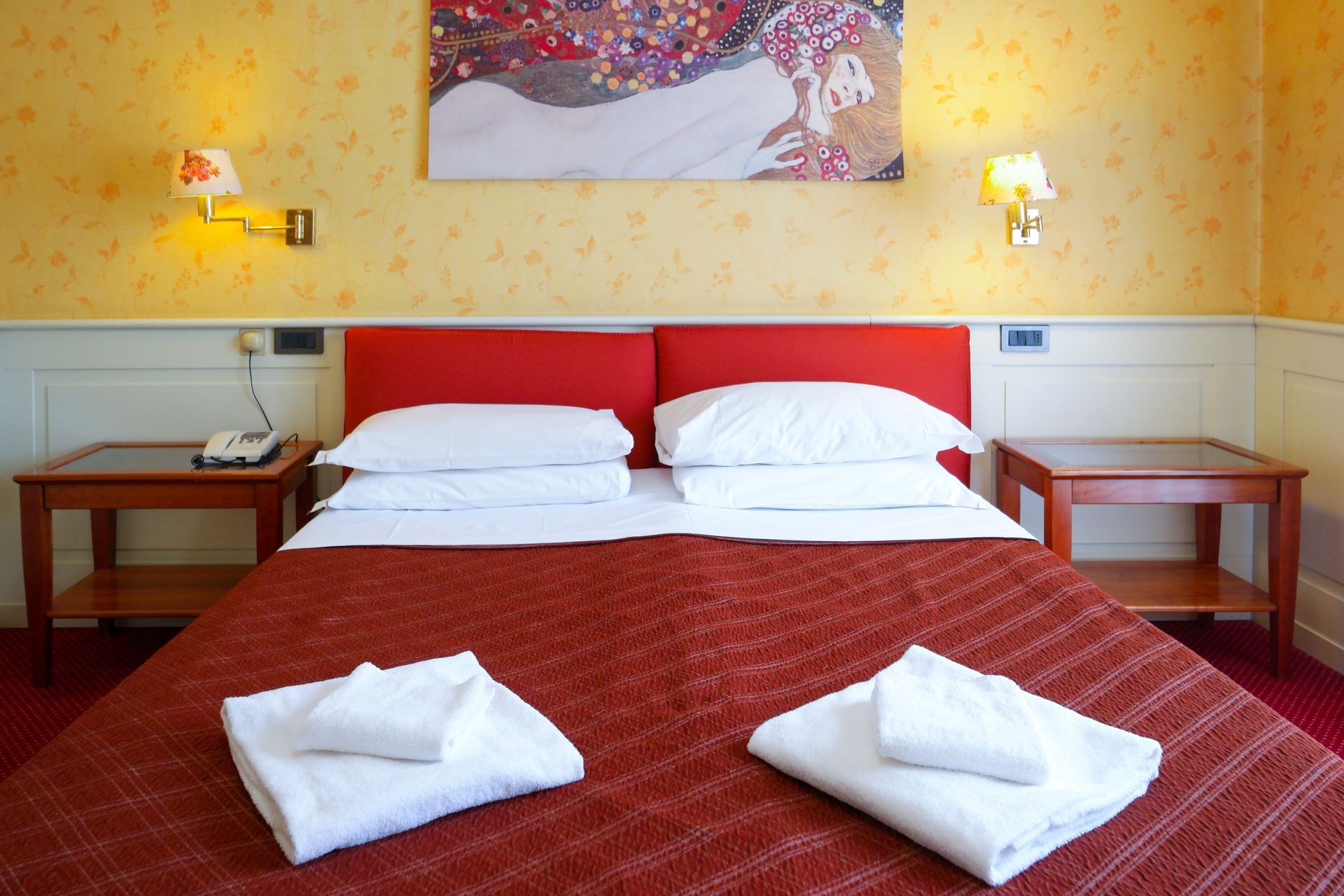 Hotel bedroom with a red bed, white linens, and two bedside tables. Yellow patterned wallpaper and artwork on the wall.