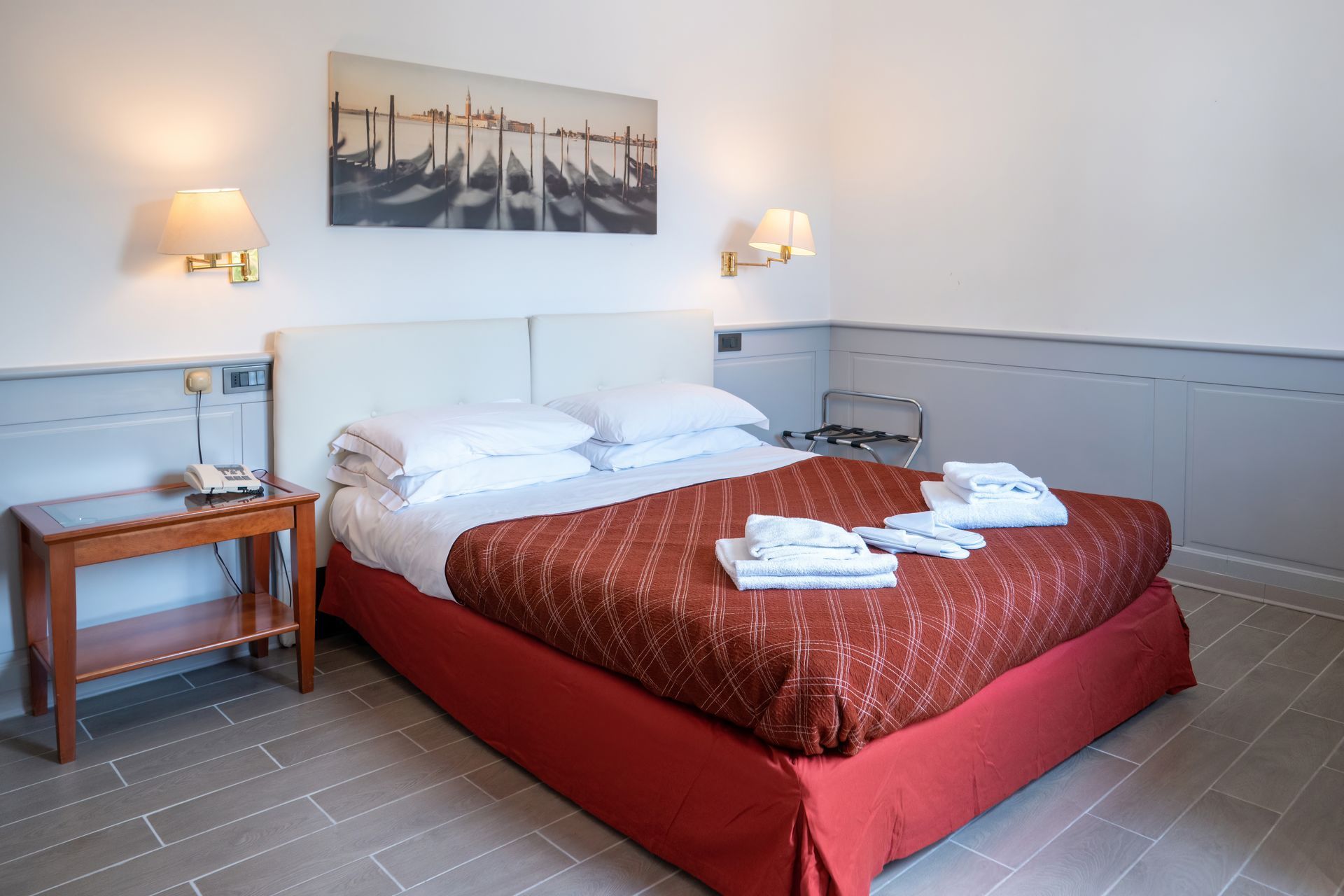 A hotel room with a made bed, nightstand, and wall art. The bed has a red cover, and there's a luggage rack in the corner.