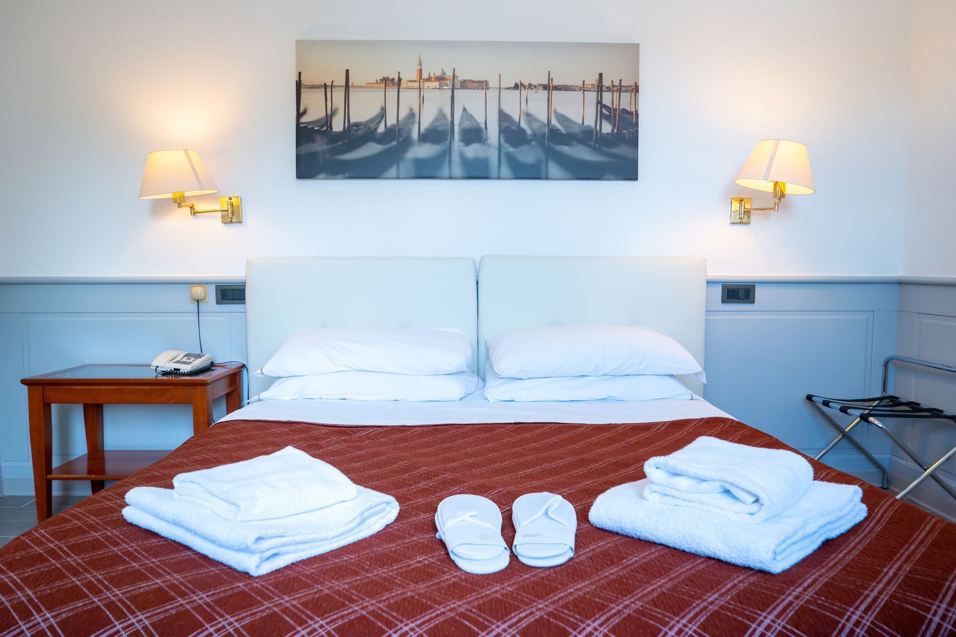 Hotel room with a made bed, towels, and slippers. A painting of gondolas hangs above the headboard.