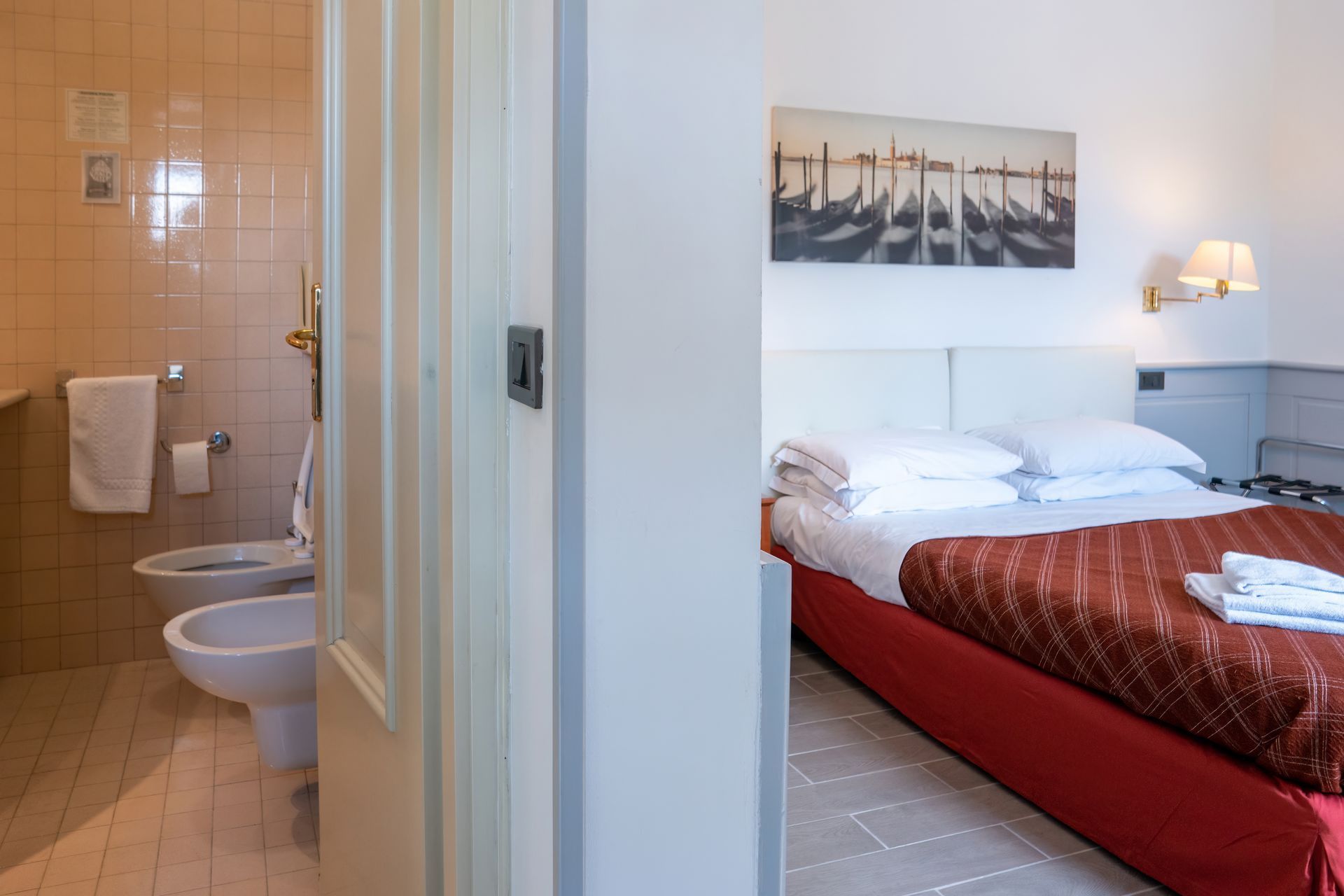Hotel room with a partially visible bathroom. Red bed, white linens, and a painting of gondolas in Venice.