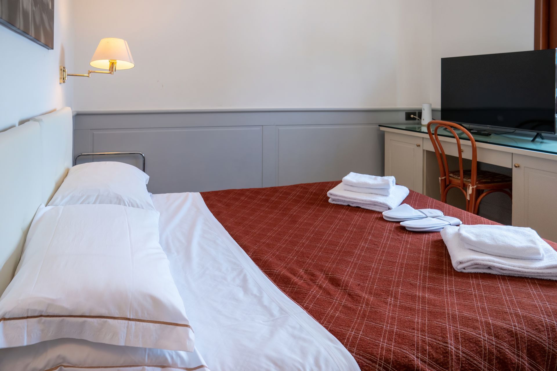 A hotel room with a made bed, a TV, a desk, and stacked white towels. The bed has a red patterned cover.