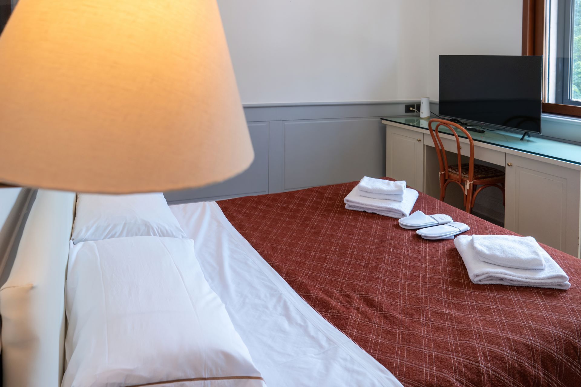 Bedroom with a bed, lamp, desk, and TV. White bedding, maroon bedspread, and towels arranged on the bed.