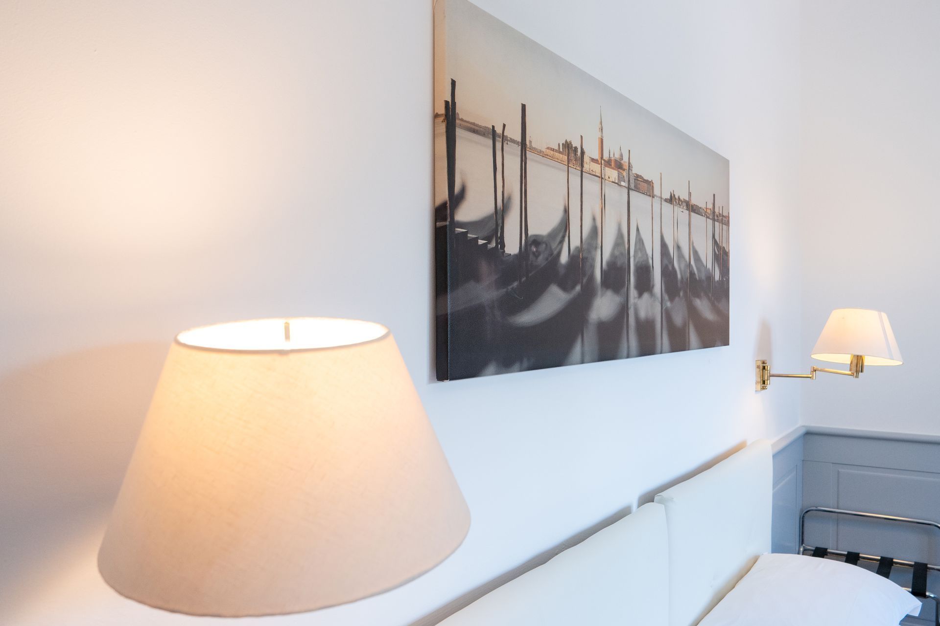 Bedroom corner with a canvas print of gondolas, a bedside lamp, and a wall-mounted lamp. White walls, neutral tones.