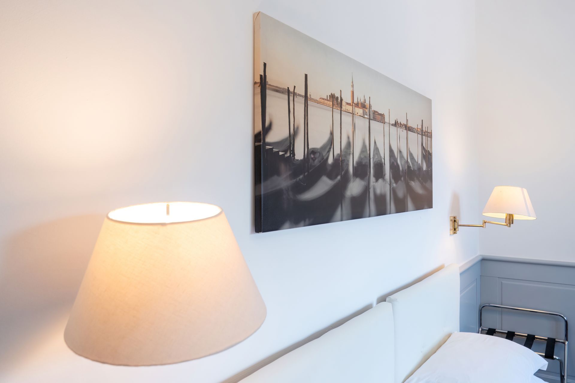 Bedroom corner featuring a white wall with a large canvas of gondolas, two lamps with cream shades, and a white headboard.