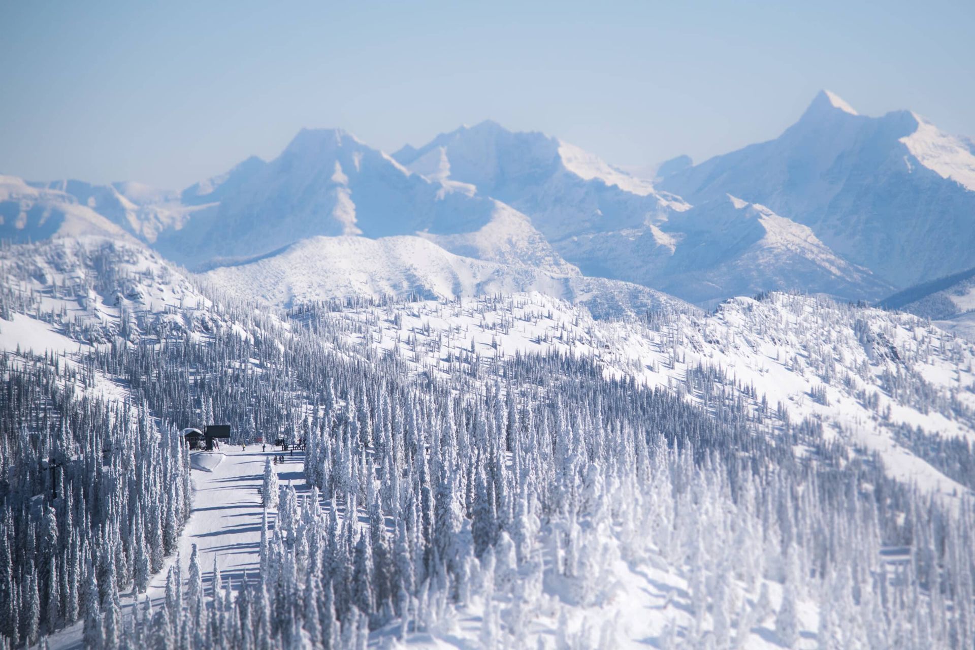 Snow-covered mountains and evergreen trees under a bright blue sky. A ski resort with buildings and ski trails is visible.