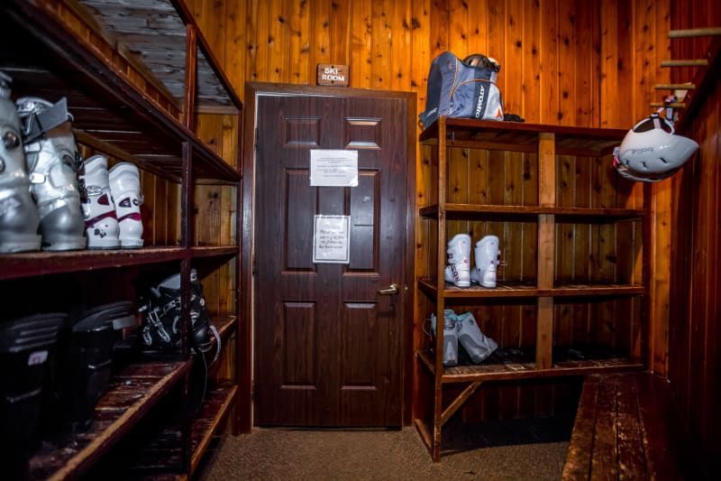 A ski boot storage room with wooden shelves and walls. Ski boots and a helmet are visible. A closed door is in the center.