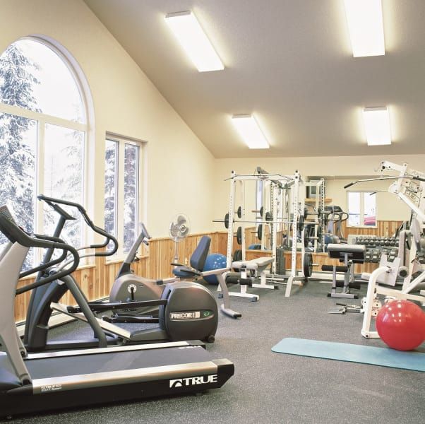 Gym with exercise equipment: treadmill, elliptical, weight machines, and exercise ball. Windows with snowy view, beige walls.