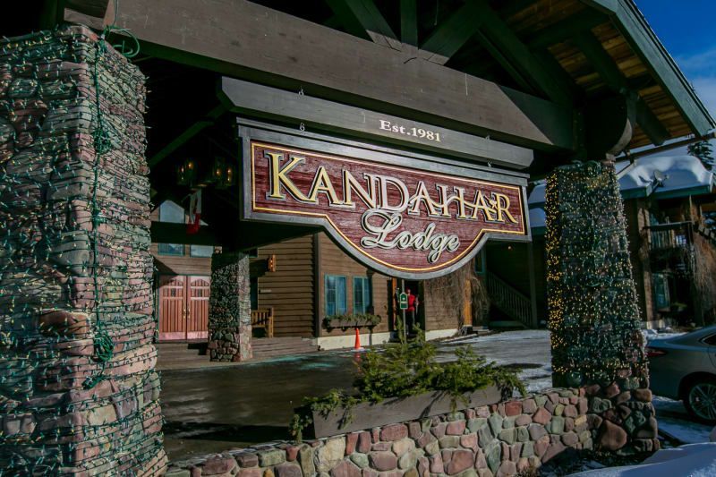 Entrance to the Kandahar Lodge with a sign under a wooden overhang. The structure is made of stone and brown wood.