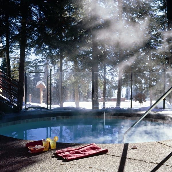 A steamy outdoor hot tub surrounded by trees in a snowy forest. A red towel and drinks sit on the edge.