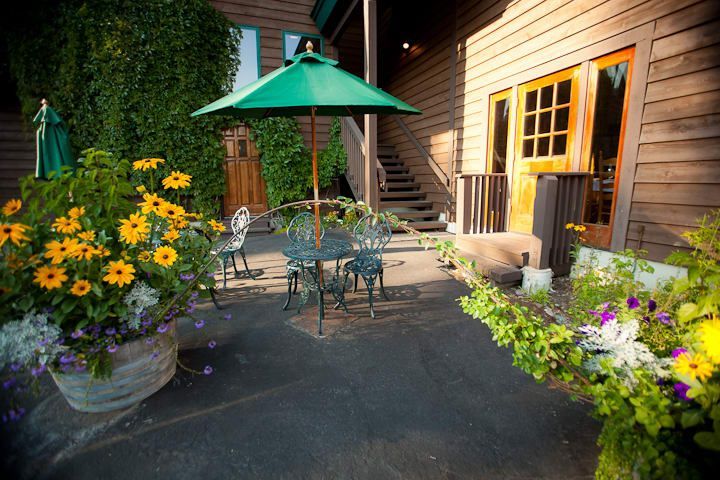 Outdoor dining area with metal furniture, a green umbrella, and flower planters next to a wooden building.