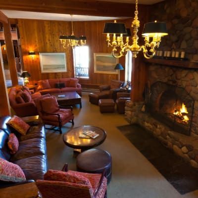 Cozy lodge living room with a stone fireplace, leather sofas, and wood paneling.  A chandelier and a lit fire create a warm atmosphere.