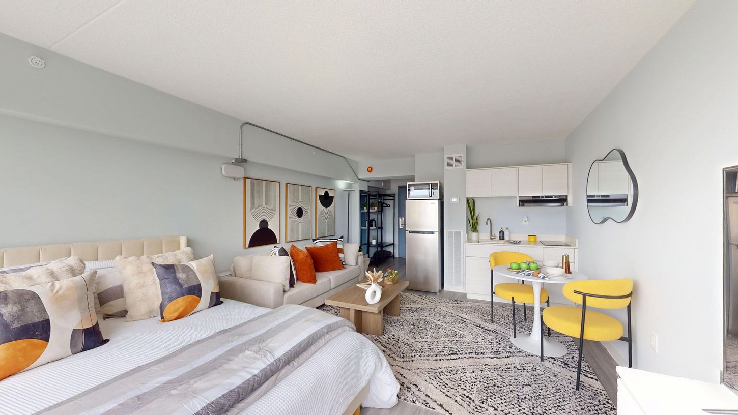 Studio apartment with bed, sofa, small kitchen, and table with two yellow chairs.