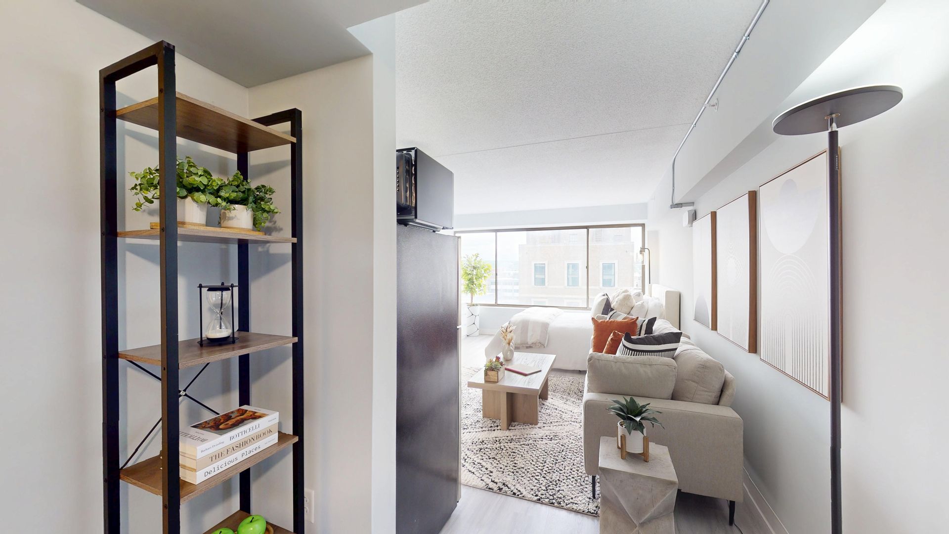 A narrow apartment hallway with a bookshelf, entering into a brightly lit living room with a sofa and windows.