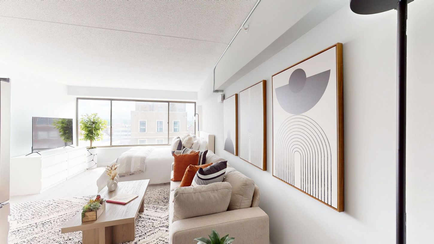 Bright living room with a white couch, orange pillows, art prints on the wall, and a large window.