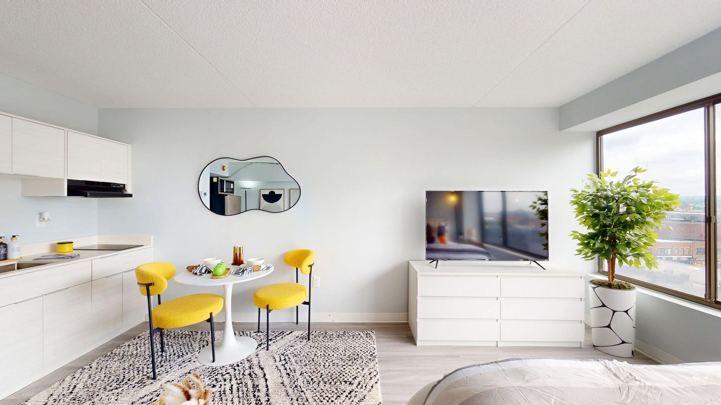 Bright studio apartment with white furniture, yellow chairs, a decorative mirror, and a large window.