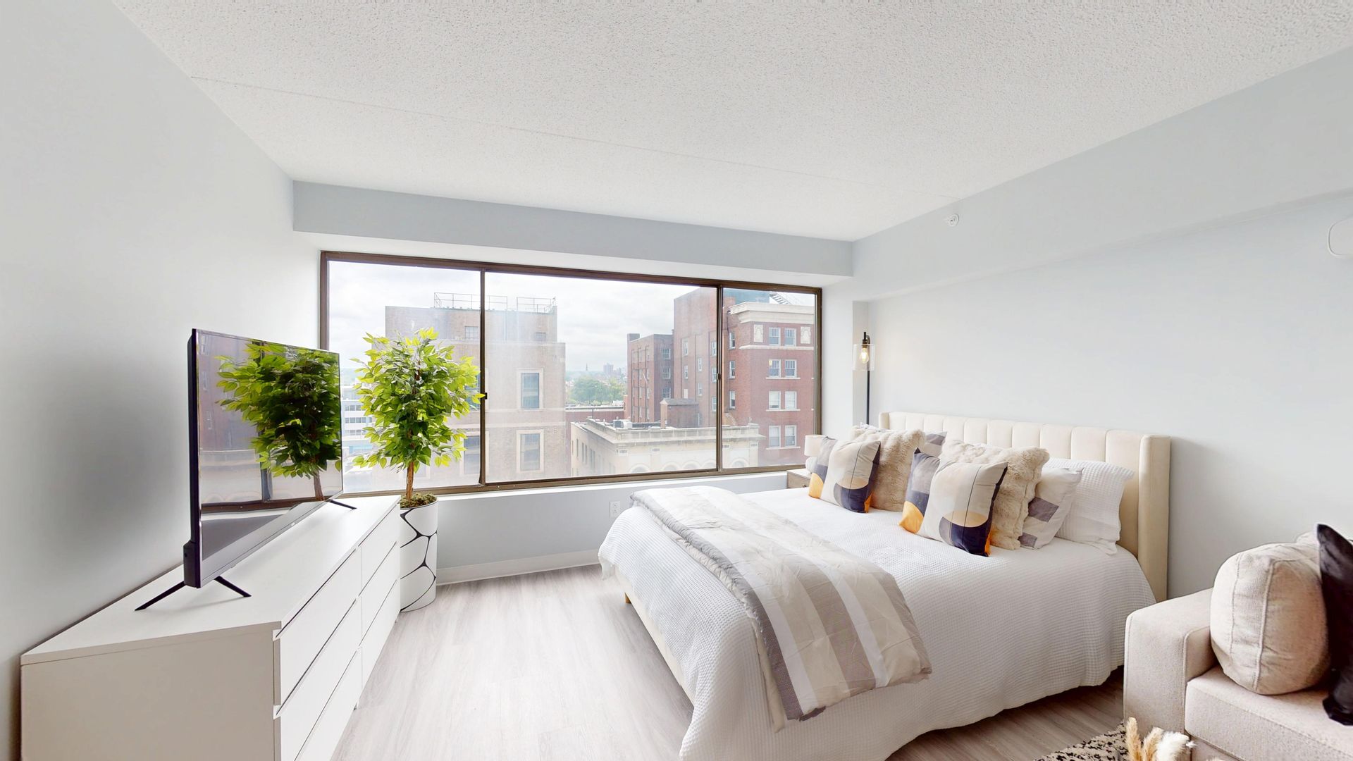 Bedroom with large windows, bed, TV, and city view. Bright, modern decor with neutral tones.