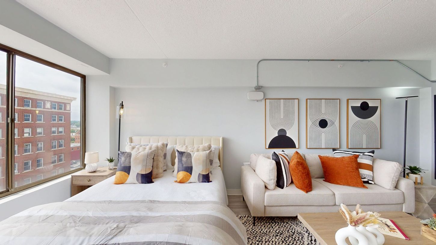 A bright, modern studio apartment with large windows, a bed, sofa, and abstract art.