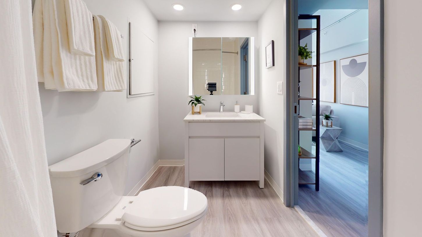 Bathroom interior with white toilet, vanity, towels, and view into the bedroom.