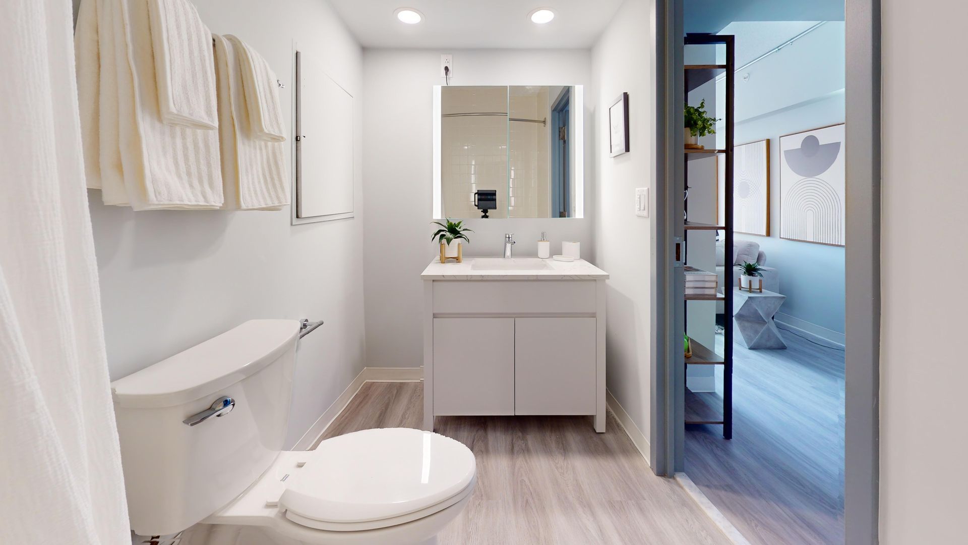 Bathroom interior with white toilet, vanity, towels, and view into the bedroom.