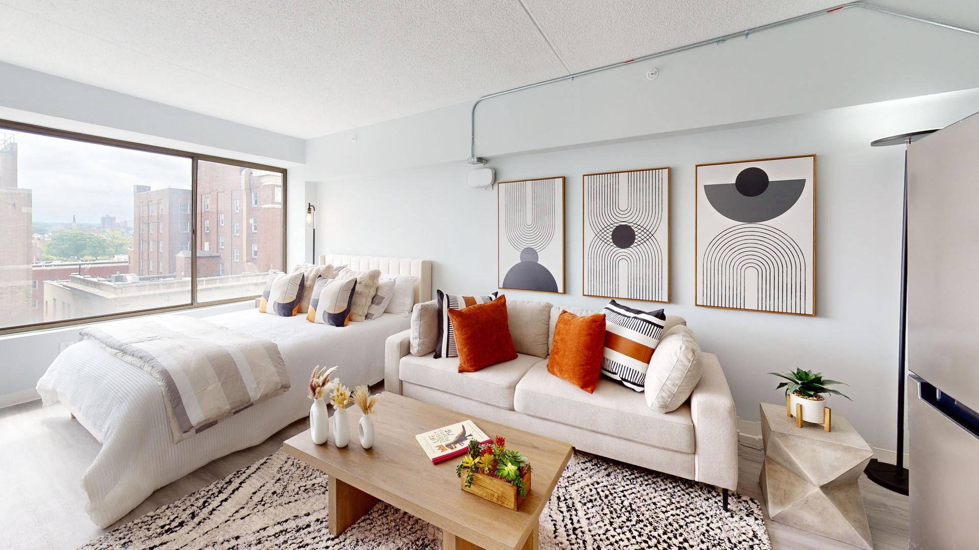 Cozy studio apartment with a bed, sofa, coffee table, rug, and artwork. Natural light and neutral colors.