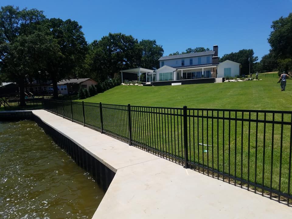 A large house sits behind a fence next to a body of water