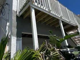 Exterior view of a building with a white deck, wooden beams, and gray stucco. Palm fronds are in front.