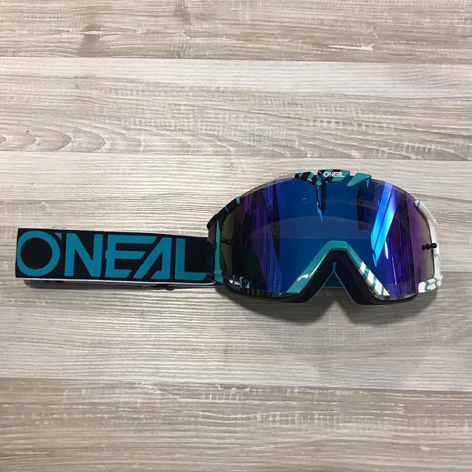 Gafas de motocross O'Neal con lente de espejo azul, correa negra y verde azulado, apoyadas sobre madera.