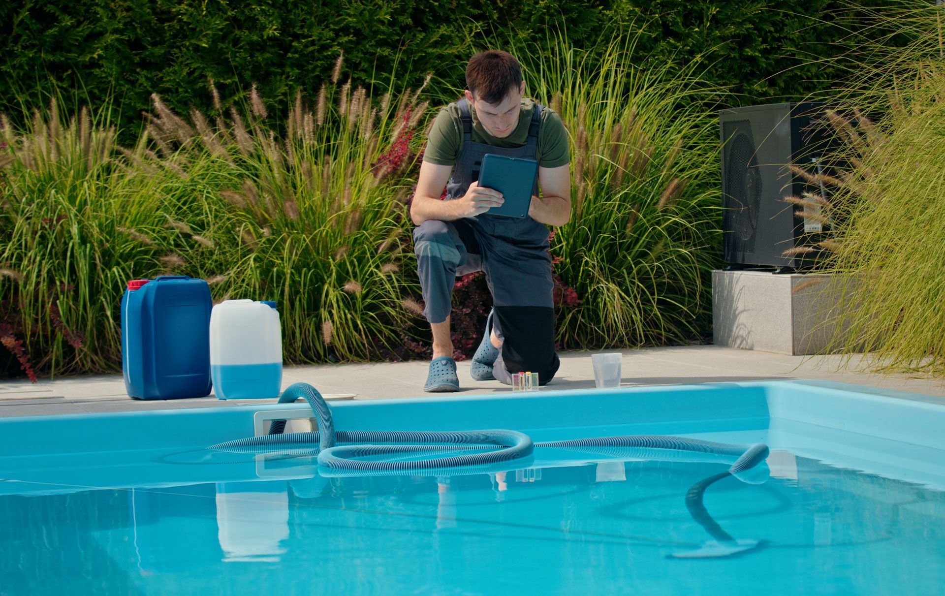 A man is kneeling next to a swimming pool looking at a tablet.