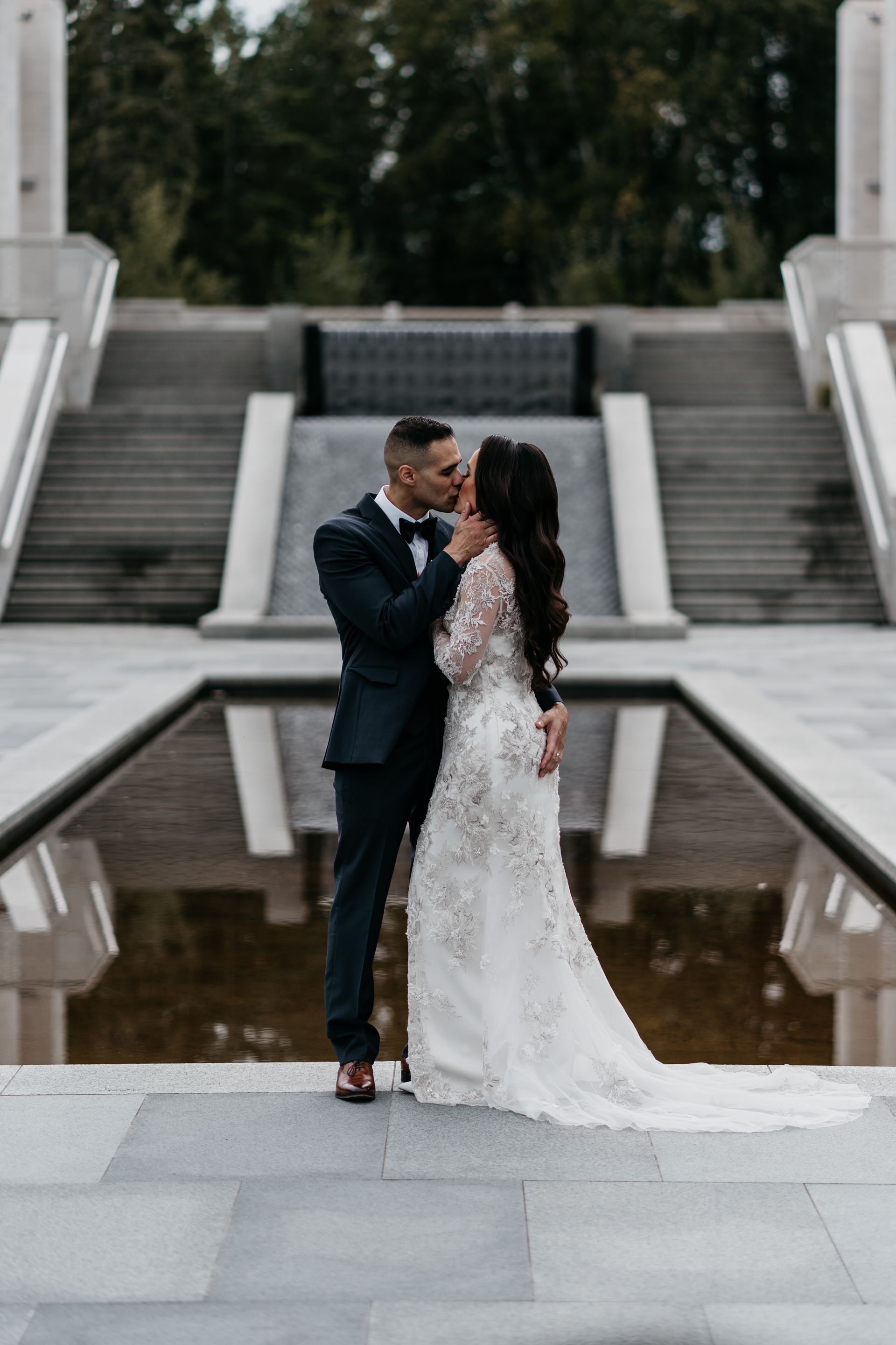 A bride and groom are kissing in front of a fountain.