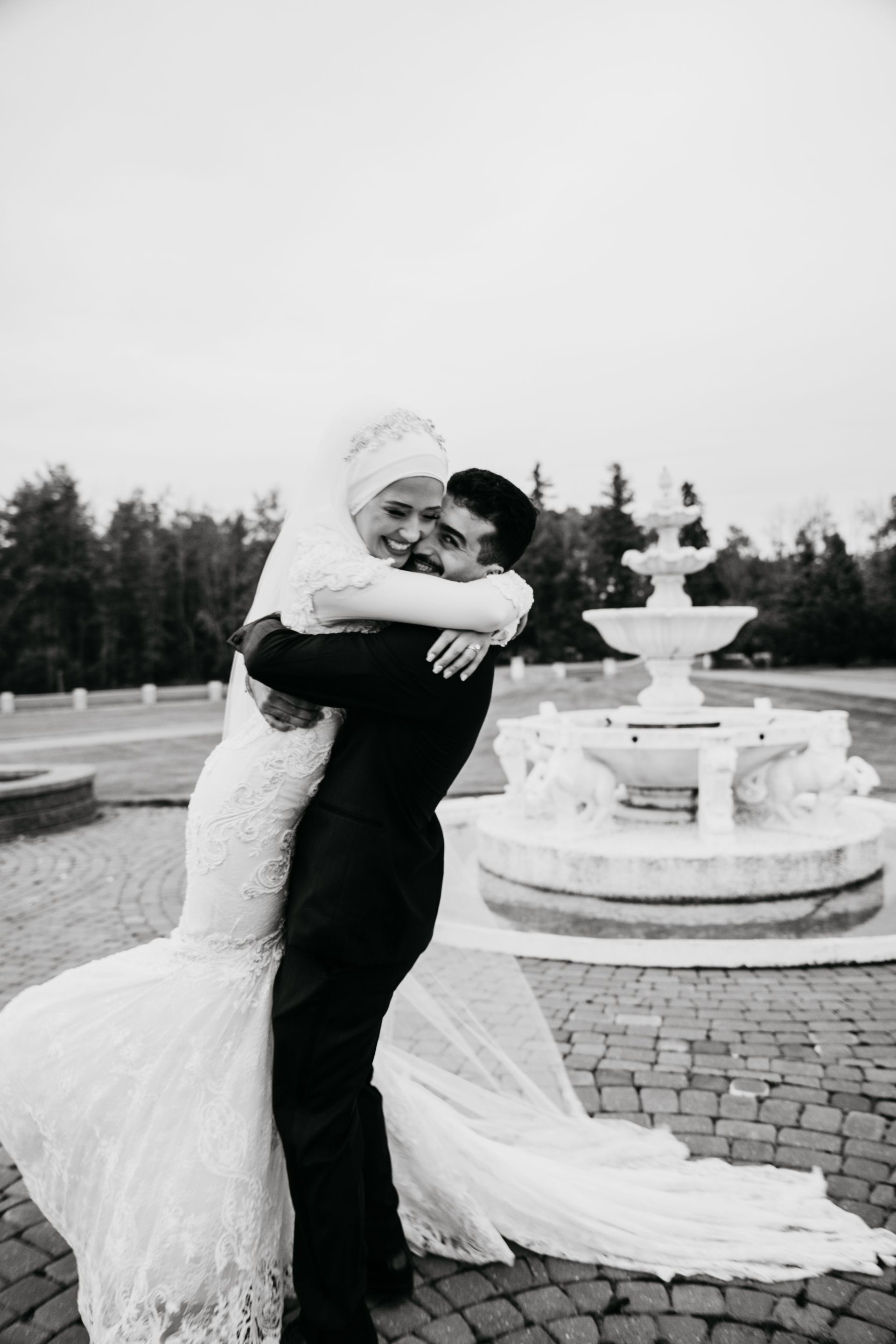 A black and white photo of a bride and groom hugging in front of a fountain.