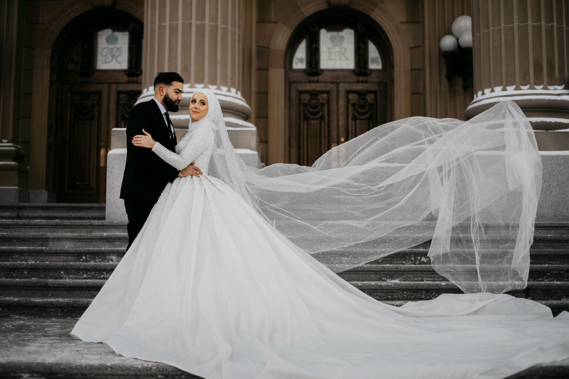 A bride and groom are posing for a picture in front of a building with their veil blowing in the wind.
