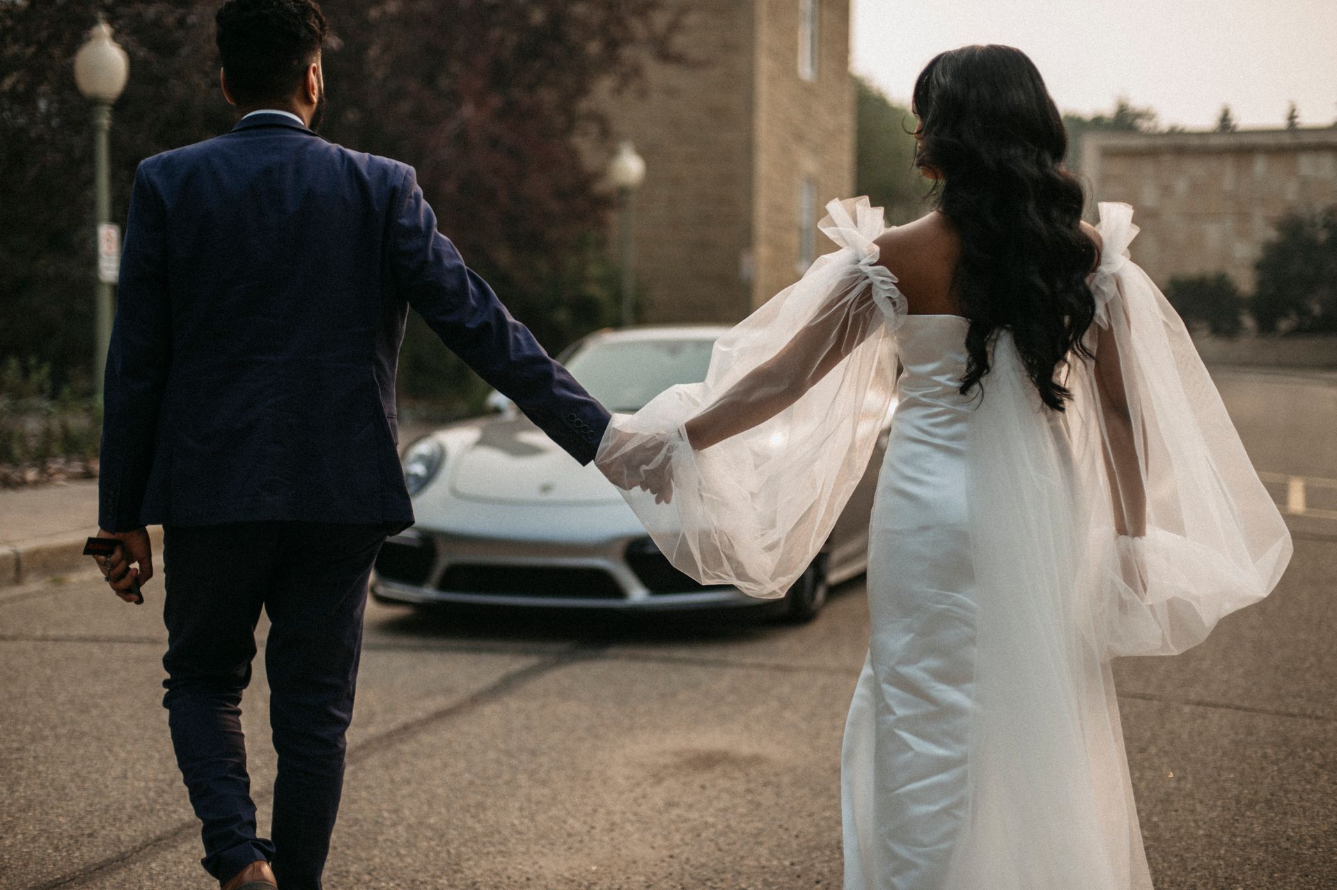 A bride and groom are walking down the street holding hands in front of a car.