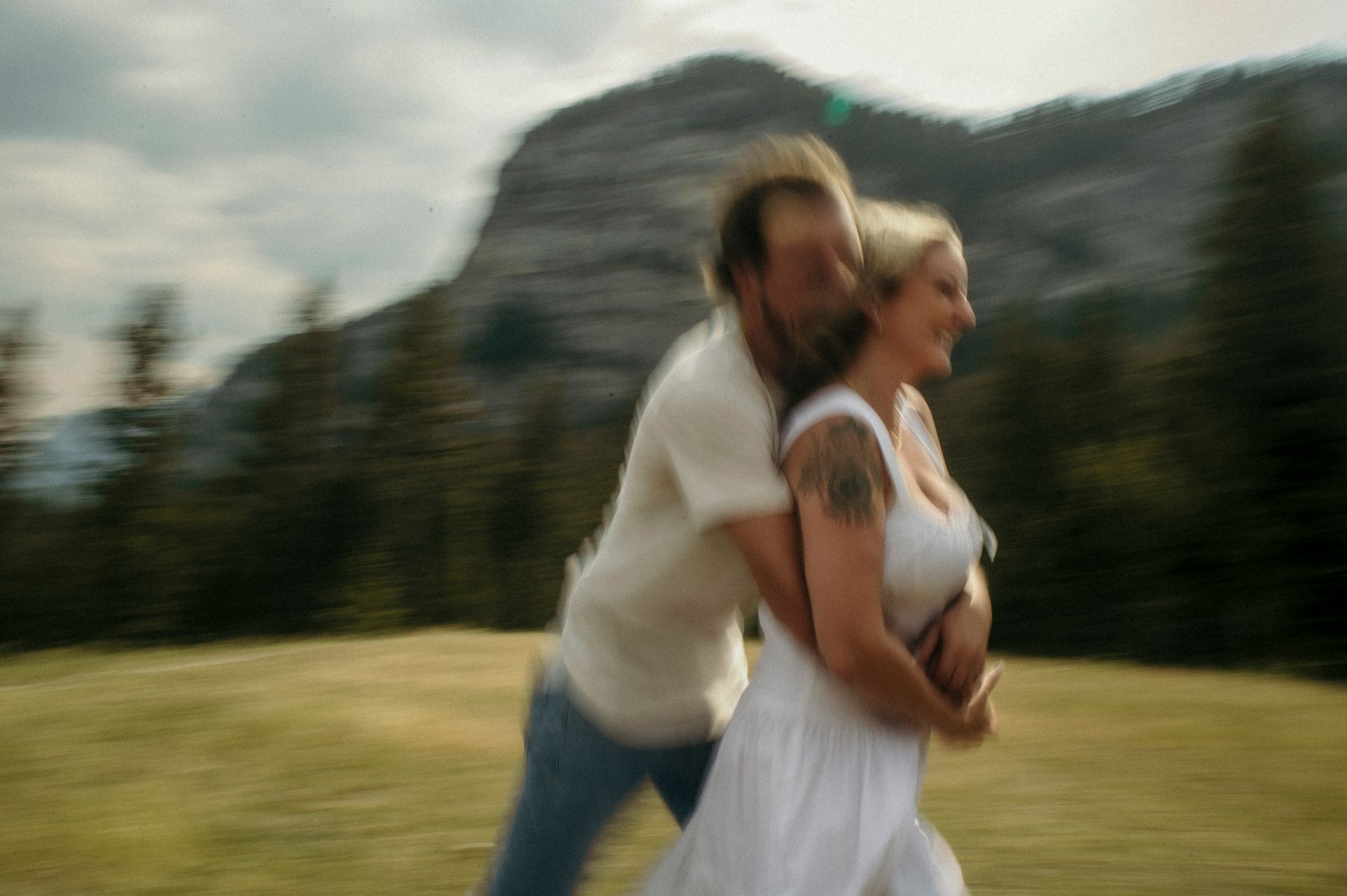 A man is hugging a woman in a field with mountains in the background.